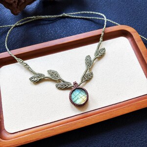 Puede incluir: Un collar de macram&eacute; con un colgante de piedra labradorita redonda con destellos iridiscentes azules y amarillos. El cord&oacute;n est&aacute; hecho de hilo verde salvia, incorporando una serie de detalles tejidos en forma de hoja a lo largo de la cadena.