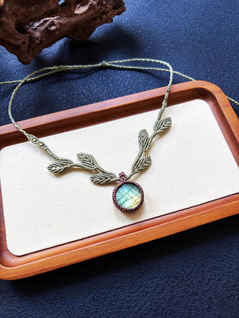 Puede incluir: Un collar de macram&eacute; con un colgante de piedra labradorita redonda con destellos iridiscentes azules y amarillos. El cord&oacute;n est&aacute; hecho de hilo verde salvia, incorporando una serie de detalles tejidos en forma de hoja a lo largo de la cadena.