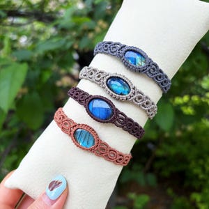 May include: Four macrame bracelets with oval blue gemstones are shown on a white cushion. The bracelets are in shades of grey, tan, brown, and rust. The gemstones have a blue and green iridescent effect.