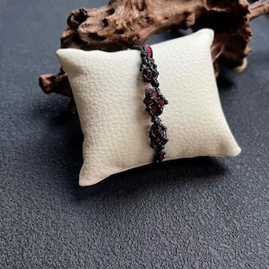 May include: A dark red gemstone bracelet with a black macrame design. The bracelet is displayed on a small, square, off-white pillow. The background is a dark, textured surface with a piece of driftwood.