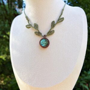 Puede incluir: Un collar de macram&eacute; con un colgante de piedra labradorita redonda con reflejos iridiscentes azules y verdes, engastado en cord&oacute;n marr&oacute;n, unido a una cadena de cord&oacute;n verde claro decorada con varios peque&ntilde;os detalles en forma de hoja.