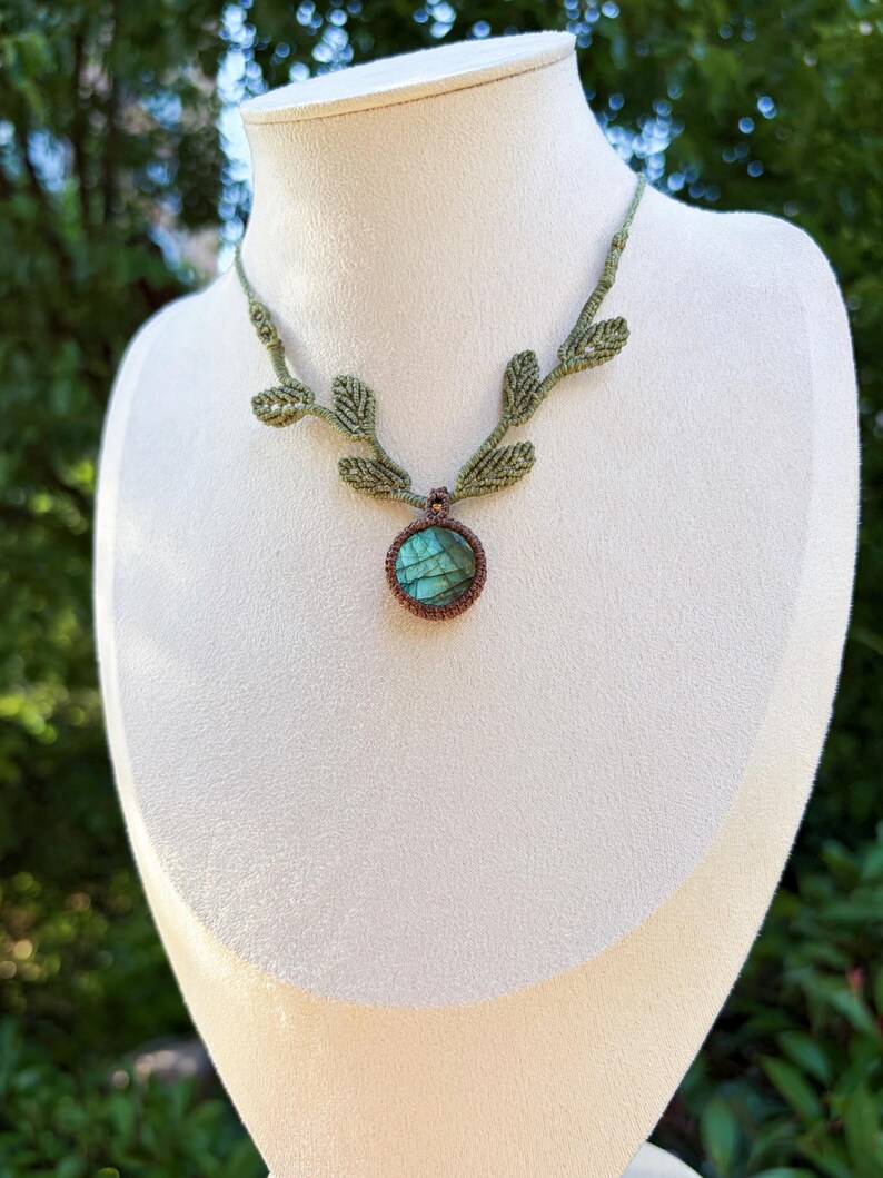 Puede incluir: Un collar de macram&eacute; con un colgante de piedra labradorita redonda con reflejos iridiscentes azules y verdes, engastado en cord&oacute;n marr&oacute;n, unido a una cadena de cord&oacute;n verde claro decorada con varios peque&ntilde;os detalles en forma de hoja.