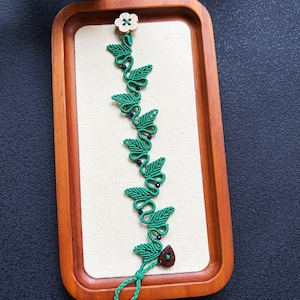 Peut inclure: Un ornement en macramé en forme de vigne fait de corde verte, composé d'une série de nœuds en forme de feuilles, de petites perles sombres, d'un bouton en forme de fleur claire en haut et d'une perle en bois sombre en forme de goutte.