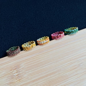 Può includere: Cinque anelli colorati, intrecciati in modo intricato, in fila. Gli anelli sono nelle tonalità di verde, giallo, arancione, rosa e verde. Sono su una superficie di legno chiaro con uno sfondo blu scuro.