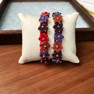 Puede incluir: Dos pulseras con diseños florales. Una es roja con flores naranjas, moradas y marrones. La otra es morada con flores naranjas y marrones. Ambas pulseras se exhiben sobre un cojín blanco.