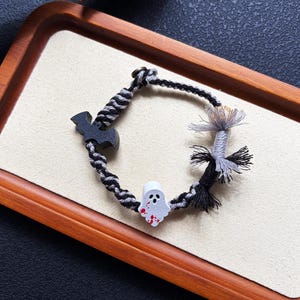 May include: A braided bracelet featuring a black and grey twisted cord, a black wooden bat charm, a white ghost charm with red paint splatters, and a decorative tassel made of grey and black threads secured with a small metal bead closure.