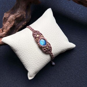 May include: A macrame bracelet featuring a central blue gemstone and gold-coloured beads. The bracelet is displayed on a white, textured cushion against a dark blue backdrop. The macrame cord is a muted brown colour.