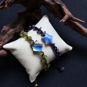 May include: Two handmade bracelets with star-shaped gemstone pendants rest on a white pillow. One bracelet features a blue star, the other a green star. Both have beaded accents and macrame cords. The background is dark.