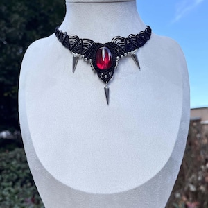 May include: Black macrame choker necklace with a large, oval, red gemstone center and silver-toned spike accents. The necklace is displayed on a white mannequin bust against a backdrop of greenery and a blue sky.