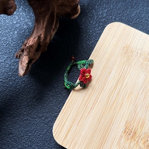 May include: A handmade ring featuring a red flower with a gold center and green leaves. The ring is placed on a wooden surface. The ring is a unique piece of jewelry.