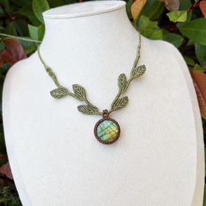 Puede incluir: Un collar de macram&eacute; con un colgante de piedra labradorita redonda en tonos azules y amarillos iridiscentes. El cord&oacute;n es de color verde oliva e incorpora detalles tejidos en forma de hoja a lo largo de la cadena.