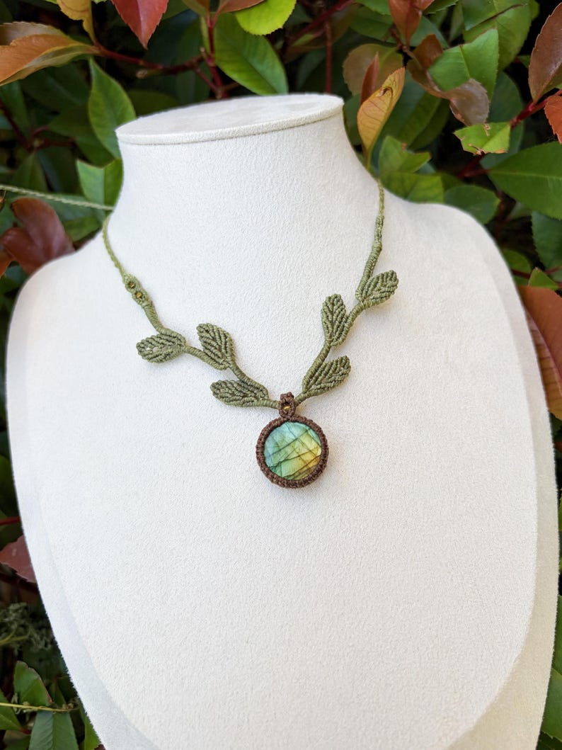 Puede incluir: Un collar de macram&eacute; con un colgante de piedra labradorita redonda en tonos azules y amarillos iridiscentes. El cord&oacute;n es de color verde oliva e incorpora detalles tejidos en forma de hoja a lo largo de la cadena.
