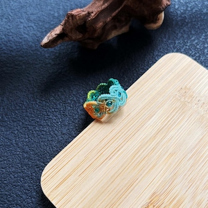 May include: A handcrafted macramé ring in teal, green, and orange hues. The ring is displayed on a light wooden surface, against a dark blue textured backdrop and a piece of driftwood.