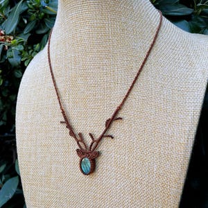 May include: A brown macrame necklace with a deer antler design. The necklace features a central pendant with a blue-green stone. The necklace is displayed on a beige mannequin.