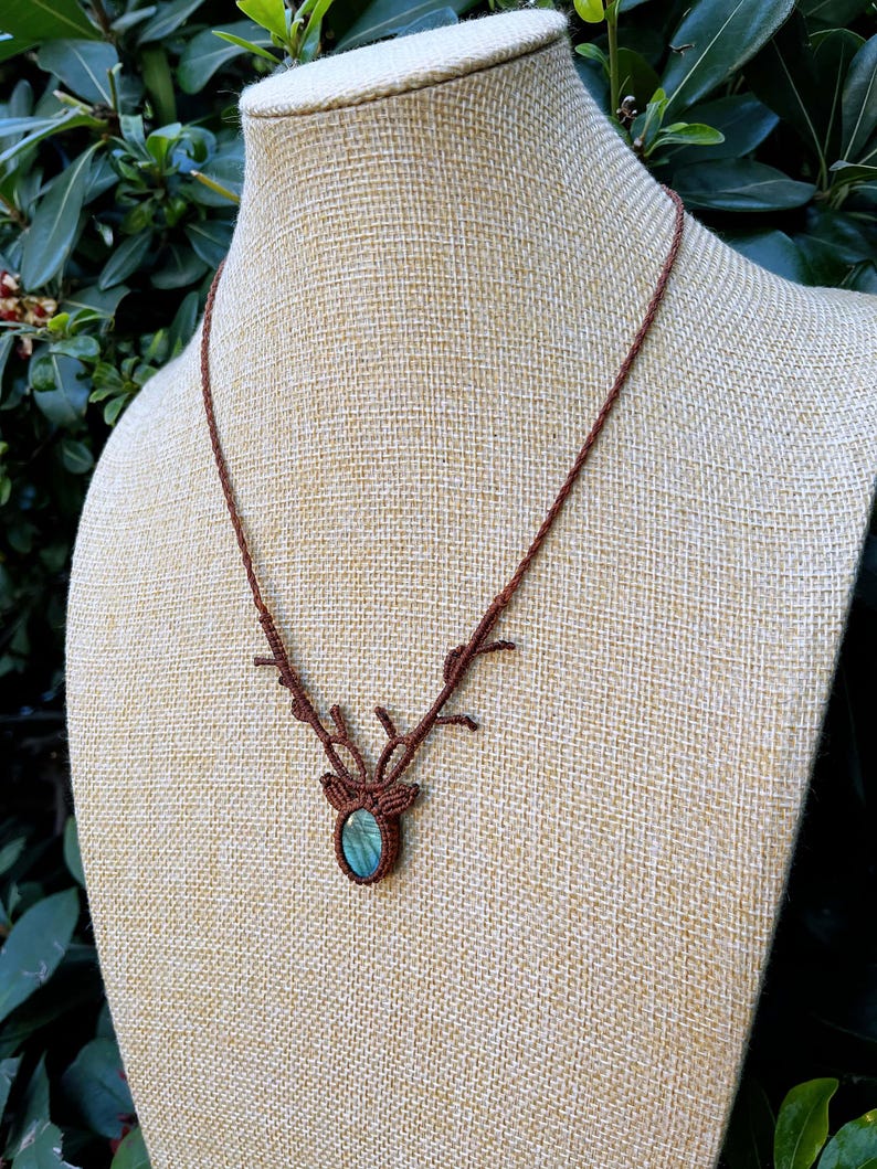 May include: A brown macrame necklace with a deer antler design. The necklace features a central pendant with a blue-green stone. The necklace is displayed on a beige mannequin.