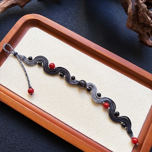 May include: A macrame bracelet featuring a wavy, braided cord that transitions from dark grey to light grey. The design is accented with round red beads, smaller black beads, and a single large, dark, spherical bead centred in the pattern.