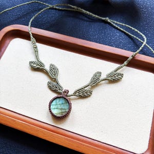 Puede incluir: Un collar de macram&eacute; con una piedra preciosa de labradorita redonda engastada en cord&oacute;n marr&oacute;n oscuro. El collar incluye detalles de cord&oacute;n verde claro con forma de hojas que se extienden desde la piedra central, unidos a una tira fina.