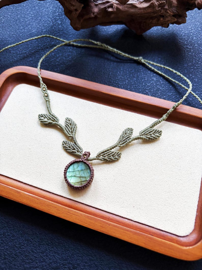 Puede incluir: Un collar de macram&eacute; con una piedra preciosa de labradorita redonda engastada en cord&oacute;n marr&oacute;n oscuro. El collar incluye detalles de cord&oacute;n verde claro con forma de hojas que se extienden desde la piedra central, unidos a una tira fina.