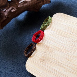 May include: Three rings crafted from woven thread in olive green, bright red, and dark brown. Each ring features a repeating geometric pattern with small yellow accents integrated into the centre of the woven design, creating a textured appearance.