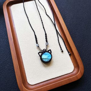 May include: A necklace featuring a round labradorite gemstone with blue iridescence, set in a black macrame frame shaped like cat ears. The cord is made of black braided thread, accented with small silver-toned beads and two translucent grey beads.