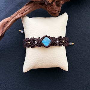May include: A brown macrame bracelet featuring a central square blue gemstone. The bracelet, with gold-coloured beads, rests on a white cushion. A piece of driftwood is in the background.