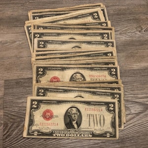 May include: A stack of vintage two-dollar bills. Each bill features a portrait of Thomas Jefferson and the text "TWO DOLLARS". The bills are in various conditions, with visible wear and tear, and are stacked on a wooden surface.