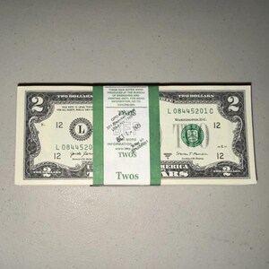 K&ouml;nnte beinhalten: Ein Stapel von Zwei-Dollar-Scheinen aus den USA, zusammengehalten von einem gr&uuml;nen Papierband. Das Band enth&auml;lt gedruckten Text, darunter 'THESE NEW NOTES WERE PRODUCED AT THE BUREAU OF ENGRAVING AND PRINTING', 'TWOS' und eine gestempelte Adresse in New York.