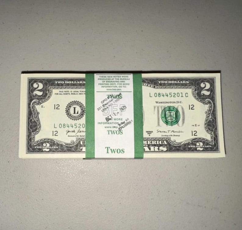 K&ouml;nnte beinhalten: Ein Stapel von Zwei-Dollar-Scheinen aus den USA, zusammengehalten von einem gr&uuml;nen Papierband. Das Band enth&auml;lt gedruckten Text, darunter 'THESE NEW NOTES WERE PRODUCED AT THE BUREAU OF ENGRAVING AND PRINTING', 'TWOS' und eine gestempelte Adresse in New York.