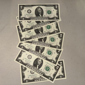 K&ouml;nnte beinhalten: Ein vertikaler Stapel von sieben Zwei-Dollar-Scheinen aus den Vereinigten Staaten, die &uuml;berlappend angeordnet sind. Jeder Schein zeigt ein Portr&auml;t von Thomas Jefferson, den Text "THE UNITED STATES OF AMERICA", "TWO DOLLARS" und fortlaufende Seriennummern.