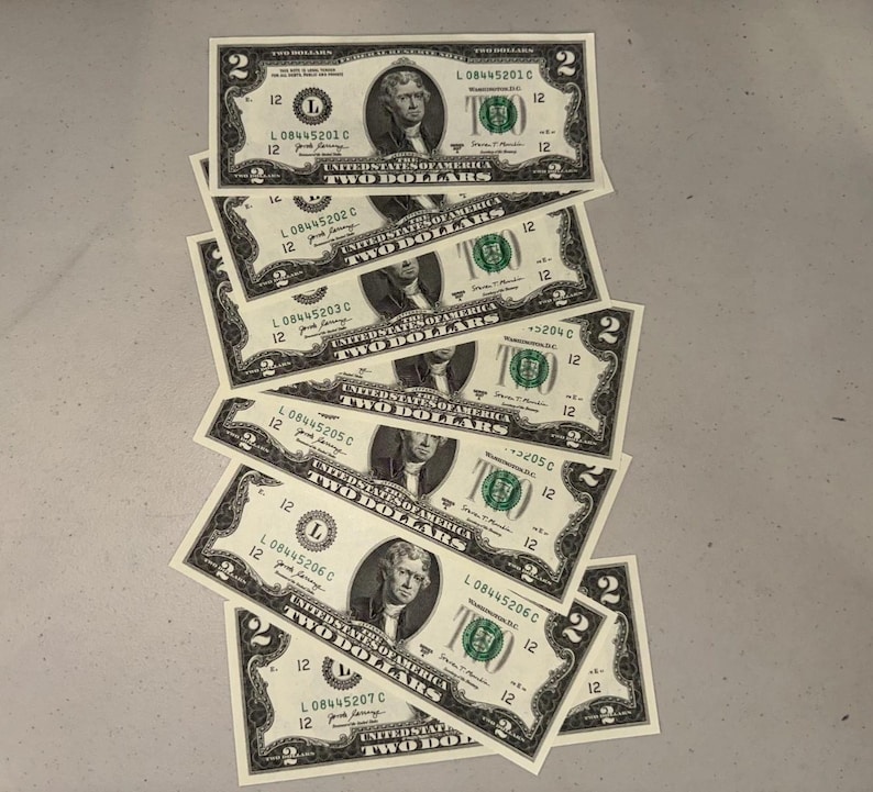 K&ouml;nnte beinhalten: Ein vertikaler Stapel von sieben Zwei-Dollar-Scheinen aus den Vereinigten Staaten, die &uuml;berlappend angeordnet sind. Jeder Schein zeigt ein Portr&auml;t von Thomas Jefferson, den Text "THE UNITED STATES OF AMERICA", "TWO DOLLARS" und fortlaufende Seriennummern.