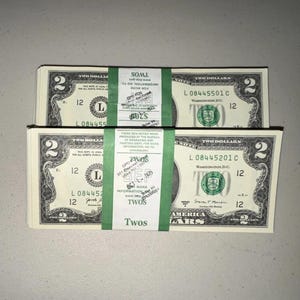K&ouml;nnte beinhalten: Zwei Stapel von US-Zwei-Dollar-Scheinen, jeweils mit einem gr&uuml;nen Papierband mit der Aufschrift TWOS versehen. Die W&auml;hrung zeigt die Zahl Zwei in den Ecken, den Text TWO DOLLARS und das gr&uuml;ne Siegel des Finanzministeriums.
