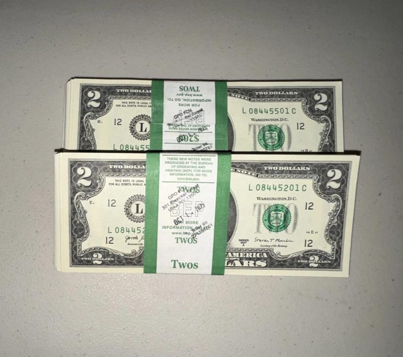 K&ouml;nnte beinhalten: Zwei Stapel von US-Zwei-Dollar-Scheinen, jeweils mit einem gr&uuml;nen Papierband mit der Aufschrift TWOS versehen. Die W&auml;hrung zeigt die Zahl Zwei in den Ecken, den Text TWO DOLLARS und das gr&uuml;ne Siegel des Finanzministeriums.
