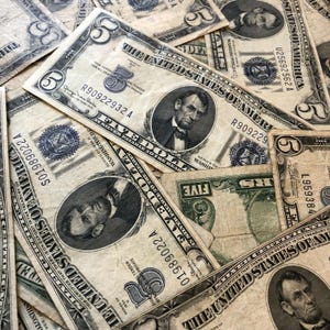 May include: A collection of vintage five-dollar bills, featuring a portrait of Abraham Lincoln. The bills are aged with visible wear and tear, showcasing the text "The United States of America" and "Five Dollars".