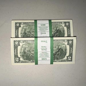 K&ouml;nnte beinhalten: Zwei Stapel von US-Zwei-Dollar-Scheinen, jeweils mit einem gr&uuml;nen Papierband versehen, das mit $200 und TWOS beschriftet ist. Die Scheine zeigen eine Gravur der Unterzeichnung der Unabh&auml;ngigkeitserkl&auml;rung und die Zahl 2 in jeder Ecke.