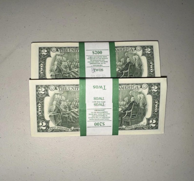 K&ouml;nnte beinhalten: Zwei Stapel von US-Zwei-Dollar-Scheinen, jeweils mit einem gr&uuml;nen Papierband versehen, das mit $200 und TWOS beschriftet ist. Die Scheine zeigen eine Gravur der Unterzeichnung der Unabh&auml;ngigkeitserkl&auml;rung und die Zahl 2 in jeder Ecke.
