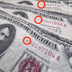 May include: Three United States five-dollar bills featuring the portrait of Abraham Lincoln. Each bill displays a red-inked star symbol before the serial number, highlighted by a red circle. The text reads United States of America and Washington D.C.