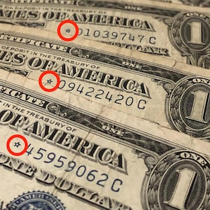 May include: Close-up of three United States one-dollar bills. The bills are slightly worn, with visible serial numbers and the words "ONE DOLLAR" prominently displayed. Each bill features a star symbol near the serial number.