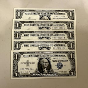 May include: Five one-dollar silver certificate bills. Each bill features a portrait of George Washington and the text "The United States of America." The bills are green and black.