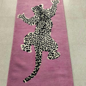 Puede incluir: Una alfombra rectangular rosa con un diseño de leopardo en blanco y negro. El leopardo está representado en una pose estilizada con los brazos levantados. La alfombra está hecha de un material suave y texturizado.