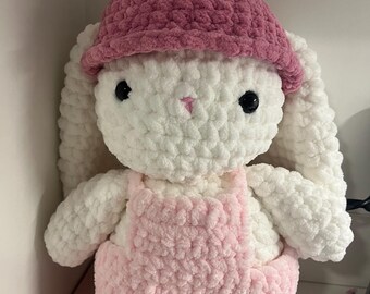 Op de afbeelding: Een handgemaakte gehaakte konijn knuffel met een witte body, roze tuinbroek en een roze hoed met een groene top. Het konijn heeft grote zwarte ogen en een kleine roze neus. De knuffel is zacht en pluizig.