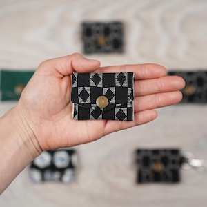 May include: A small, black and white patterned fabric wallet with a gold-colored button closure. The wallet is held in a hand, with other similar wallets in the background. The pattern is a repeating geometric design.