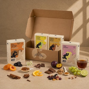 Op de afbeelding: Een kartonnen geschenkdoos met drie rechthoekige koffiedozen met de labels Bali Natural Escape, Toraja Citrus Delight, Kerinci Honey Velvet en Ijen Legacy. De display bevat gedroogde abrikozen, pruimen, koffiebonen, chocolade, pruimen, walnoten, rozijnen, kaneel, steranijs en een kopje koffie.