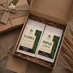 Könnte beinhalten: Eine Pappgeschenkbox mit zwei weißen Kaffeetüten der Marke STIA Coffee Roasters. Eine Tüte ist mit FLORES Nusa Tenggara Timur beschriftet, mit Noten von Orange, Mandel und Schokolade. Die andere ist mit PAPUA WAMENA beschriftet, mit blumigen, karamellisierten und Traubennoten.