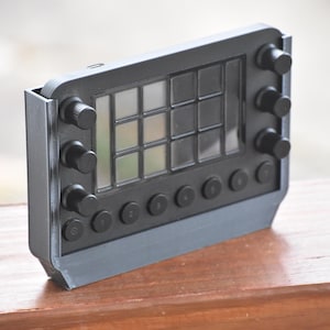 May include: A black electronic device with a grid of clear squares and several black knobs and buttons. The device is rectangular with a gray frame and is resting on a wooden surface.