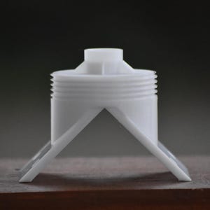 May include: A white plastic component with a cylindrical body and a threaded section. It has a small, raised top and two angled support legs. The object is designed for a specific function, likely in a larger assembly.