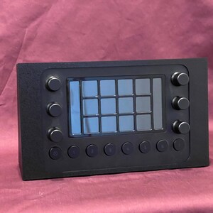 May include: A black electronic device with a grid of 16 square buttons in the center, surrounded by several round knobs and buttons. The device has a rectangular shape and is likely used for audio or video production.