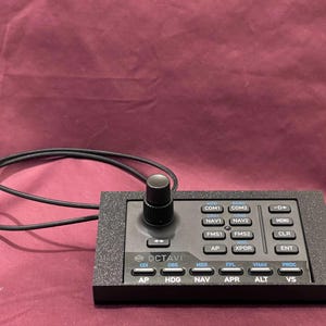 May include: A black control panel with multiple buttons and a rotary knob. The panel has labels such as "COM1", "NAV1", "AP", and "VS". A black cable extends from the panel. The background is a maroon fabric.