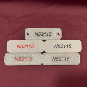 May include: Several rectangular, off-white tags with rounded corners. Each tag features the number "N52115" in black or red. One tag has a hole. The tags are displayed on a maroon background.