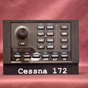 May include: A black and gray aircraft instrument panel with multiple buttons, knobs, and a digital display. The text "Cessna 172" is displayed at the bottom. The panel includes labels such as "HDG," "COM1," and "NAV1."