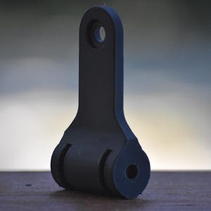 May include: A black, 3D-printed object with a rectangular top and a rounded base. The object has a hole at the top and two circular cutouts on the base. It is resting on a wooden surface.